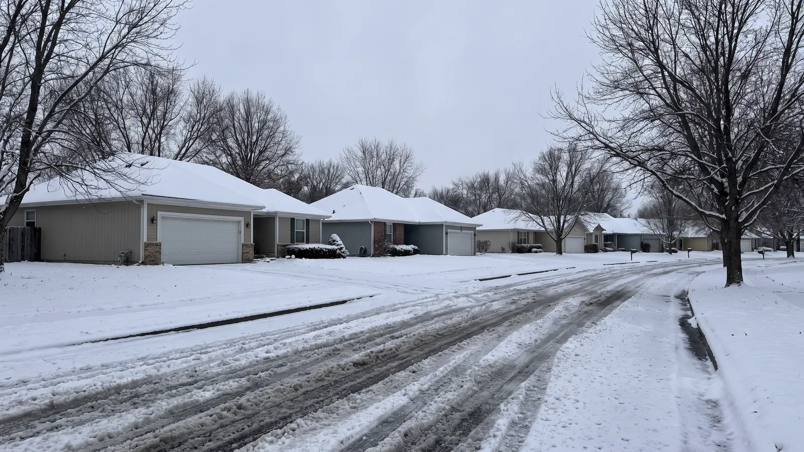 Row of suburban houses with two-car garage doors along a snow-covered Kansas street, bare trees and grey winter sky overhead